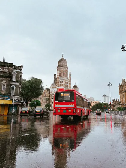 A classic red BEST bus makes its way through the rain-slicked streets, with the historic BMC building standing tall in the background. A typical, yet beautiful, Mumbai monsoon scene.