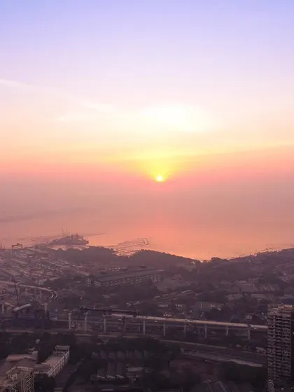 A hazy sunrise over Mumbai, with the sun appearing as a bright orb above the Bandra-Worli Sea Link and the coastline.