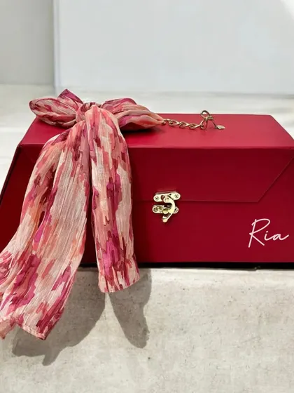 The packaging is part of the gift. This stunning red trapezoid box, personalized with the name "Ria," is elegantly tied with a printed scarf, creating a luxurious and memorable unboxing experience.