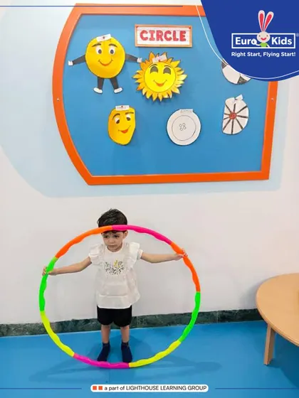 A child proudly holds up a hula hoop, surrounded by circle-themed crafts on the wall. Our themed days make abstract concepts like shapes tangible and exciting for young learners.
