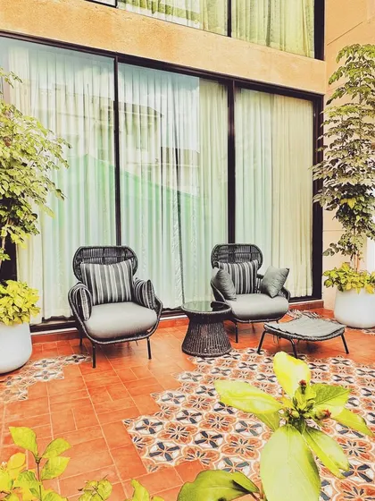 This intimate patio features comfortable armchairs set upon a beautiful floor of terracotta and patterned tiles. Tall potted plants frame the space, creating a seamless transition between the indoor and outdoor areas.