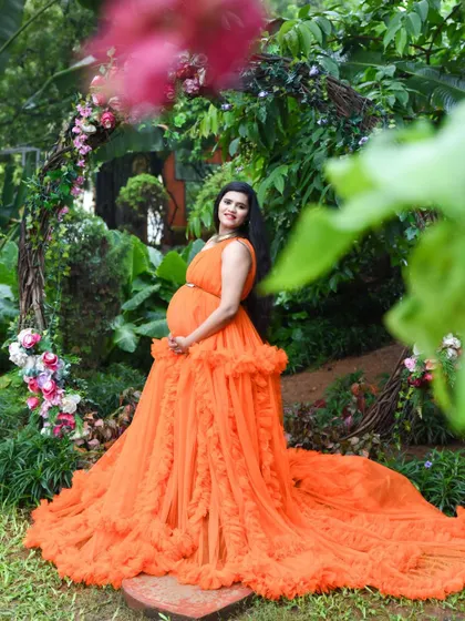 A full-length shot showcasing a magnificent orange ruffled gown in a lush garden. The mom-to-be is framed by a rustic arch, creating a fairytale-like scene.