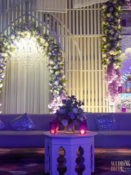 A comfortable and stylish lounge seating area. The soft lavender sofa is accented with sequined purple cushions, and the floral arrangements continue the purple and white color story.