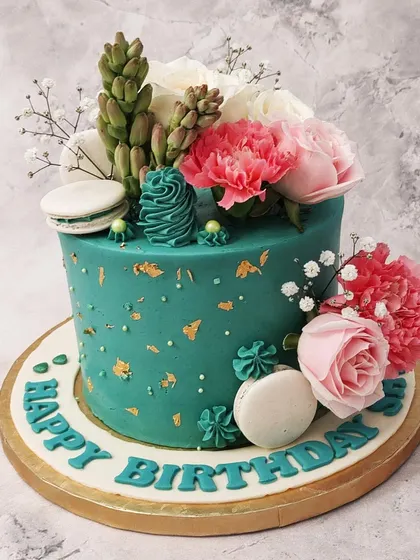 We combine the beauty of succulents and flowers in this unique green floral birthday cake. A rich teal cake is the perfect backdrop for a lush arrangement of fresh pink and white flowers, edible succulents, and macarons, creating a vibrant, botanical look.
