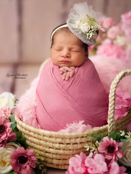 A beautiful newborn girl, wrapped in pink, rests in a woven basket surrounded by delicate pink flowers. The tiny floral fascinator adds a touch of elegance to this classic newborn pose.