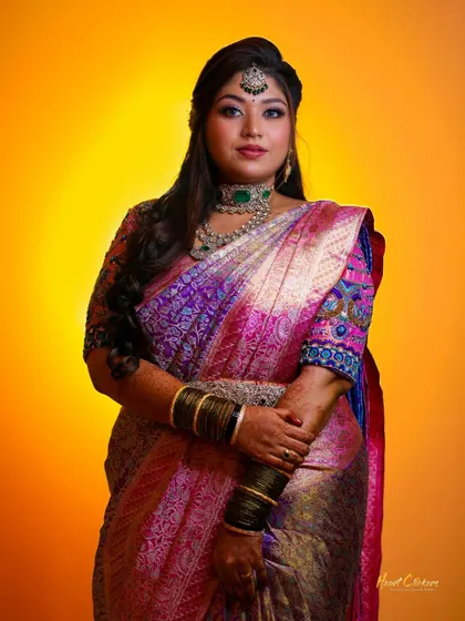 A full-length bridal portrait showcasing a magnificent purple and pink silk saree against a warm, glowing background.