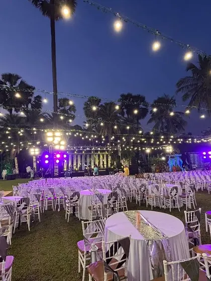 An outdoor reception setup at dusk with purple uplighting. The combination of string lights, elegant table settings, and ambient lighting creates a sophisticated and magical atmosphere.
