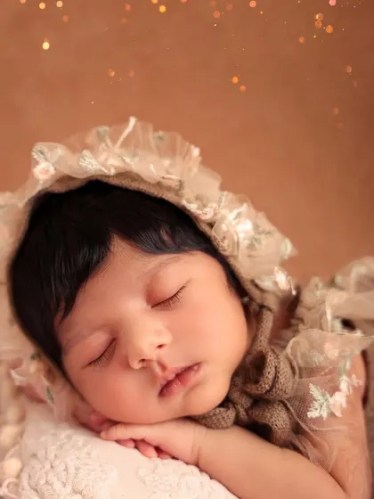 A close-up portrait of a sleeping newborn wearing an elegant, vintage-style lace bonnet. The soft focus and warm tones give this image a timeless, heirloom quality.