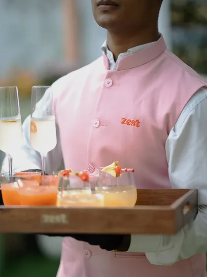 A server in a sharp pink "Zest" uniform holds a tray of welcome drinks.