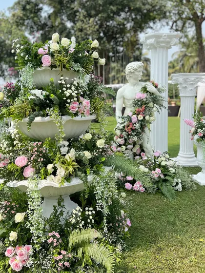 A tiered fountain and a classic Roman-style statue, both lavishly decorated with flowers, adding a touch of European elegance to the Goan coastline.