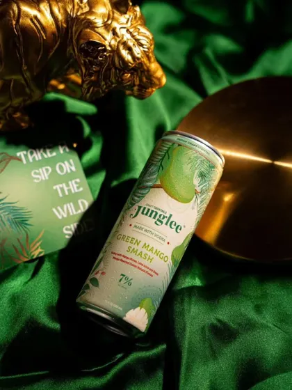 A styled product shot of Junglee's Green Mango Smash. The use of green satin fabric and gold props creates a luxurious and tropical vibe.