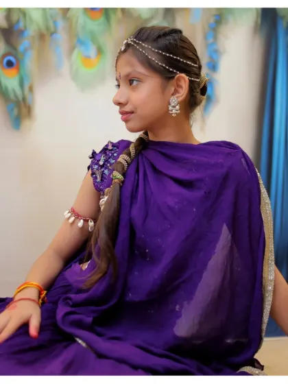 A thoughtful profile shot of a girl dressed as Radha, highlighting the traditional jewelry and braided hairstyle against a peacock feather backdrop.