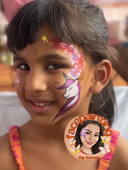 A smiling girl proudly showing off her unicorn and floral face paint. The red glitter adds a beautiful pop of color.