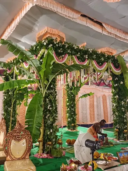 A traditional South Indian mandap setup inside a hall, featuring pillars wrapped in greenery and banana plants.