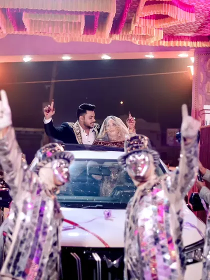 A high-energy wedding exit, with the couple celebrating from their car while a dance troupe in mirror-work costumes performs for them.