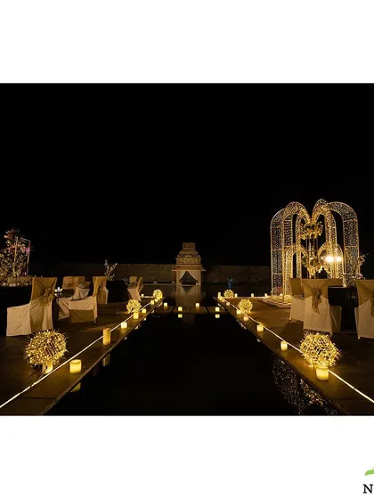 A romantic outdoor Sangeet setup at night. The area is lit with fairy lights and candles, with illuminated heart-shaped structures creating a magical and intimate setting by the water.