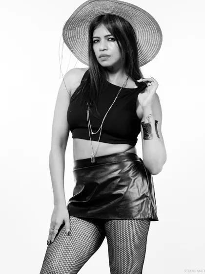 A black and white portrait of a model in a black crop top, leather skirt, and a wide-brimmed hat. The high-key lighting gives it a clean, editorial feel.