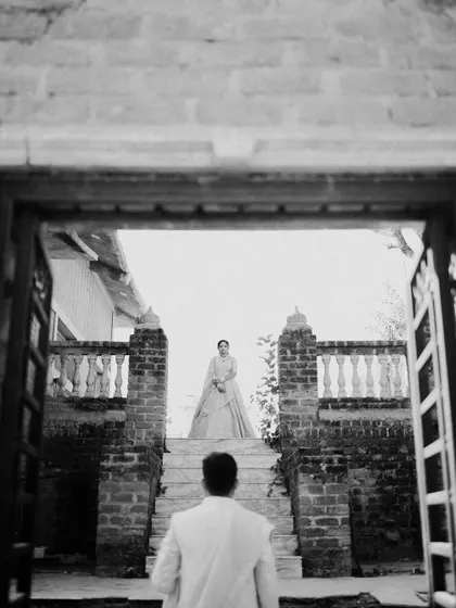 A dramatic black and white 'first look' style shot. The groom waits at the bottom of the stairs as his bride appears, a moment full of anticipation and romance, framed beautifully by the doorway.