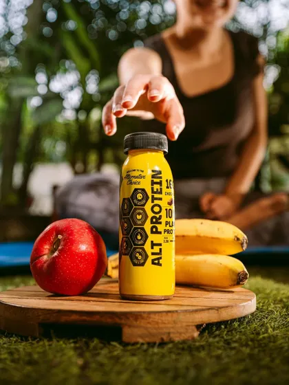 A post-yoga lifestyle shot with Alt Co.'s protein drink. A model in a yoga pose reaches for the bottle, clearly linking the product to wellness, fitness, and recovery.