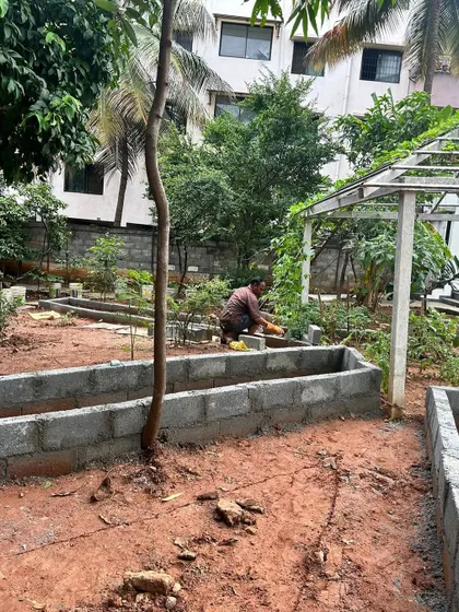 The construction phase of a large vegetable garden. A team member is working on the raised beds made of concrete blocks, with a pergola frame visible in the background.