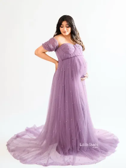 A full-length portrait of an expecting mother in a delicate lavender gown adorned with pearls. She stands gracefully against a white background, showcasing the elegant design of the dress.
