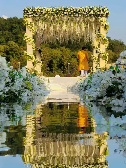 A stunning reflection shot of the all-white floral mandap on a mirrored aisle, doubling the beauty of this serene riverside setup.