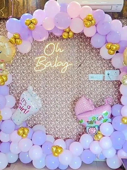 A close-up of the lavender and pink "Oh Baby" shower decoration. The neon sign glows warmly against the patterned wallpaper, and the themed foil balloons add a playful touch.