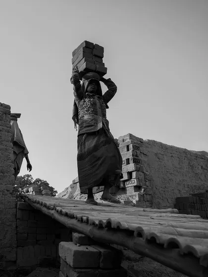A woman carefully walks across a narrow wooden plank, balancing a heavy load of bricks on her head. The image highlights the skill and risk involved in her work.