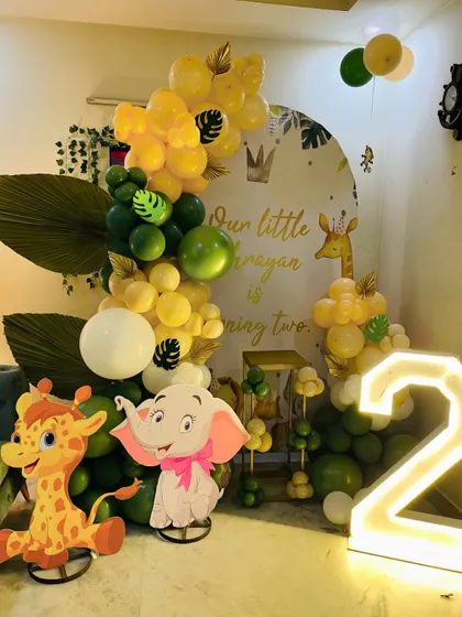 A cute jungle-themed second birthday party. This home setup includes a green and yellow balloon arrangement, character cutouts of a giraffe and elephant, and a large light-up number '2'.