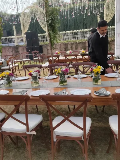 A close up of the guest dining tables at a rustic themed outdoor wedding. The wooden tables are paired with cross back chairs and simple floral centerpieces in mason jars for a charming look.