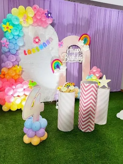 A cheerful Rainbow theme birthday decoration. This setup uses a pastel rainbow color palette for the balloon arch, with rainbow cutouts, a personalized backdrop, and themed pedestals on a green turf floor.