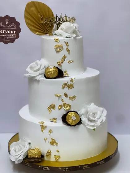 A classic three-tier wedding cake in white and gold. Adorned with Ferrero Rocher chocolates and white roses, this cake is both elegant and delicious, perfect for a sangeet or reception.
