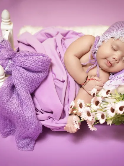 A cozy newborn portrait with the baby sleeping on a miniature bed, draped in soft lavender fabrics. The warm lighting creates a gentle and dreamy atmosphere.