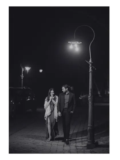A romantic black and white photo of the couple walking down a street at night, illuminated by a single lamppost. This image has a classic, old-world cinematic charm.