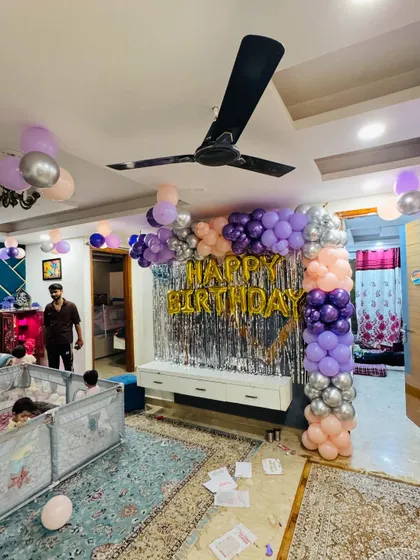 A birthday decoration in a living room with a purple, peach, and silver balloon arch over a TV unit.