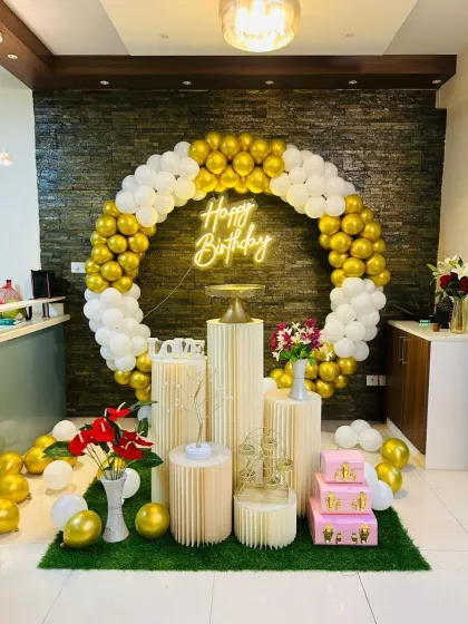 A full circle balloon frame in white and gold makes a stunning centerpiece for this birthday celebration. The setup includes a neon sign and multiple pedestals for cake and decor display.