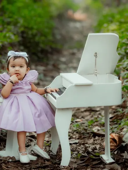 A little pianist in a beautiful lavender dress, posing with a miniature piano in a lush, green, forest-like setting.