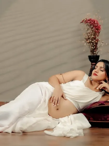 A serene portrait of the mother-to-be resting, her hand on her bump. The white saree and the soft, diffused lighting in the studio create a tranquil and dreamy mood.