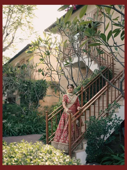 The bride poses elegantly on a staircase, her red lehenga a stunning contrast against the natural, earthy tones of the building and foliage.