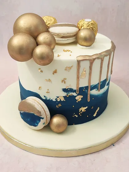 We design a cake of pure modern luxury with this blue and gold drip cake. A deep blue ombre effect is accented with gold drips, gold leaf, and an arrangement of golden spheres, Ferrero Rocher, and macarons, creating a sophisticated look for any celebration.