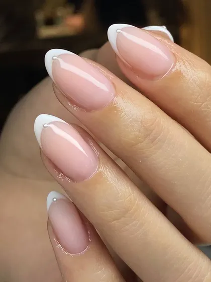 A modern twist on a classic. This elegant French manicure on almond-shaped nails is elevated with a tiny, delicate pearl accent at the base of each tip, adding a touch of sophisticated detail.