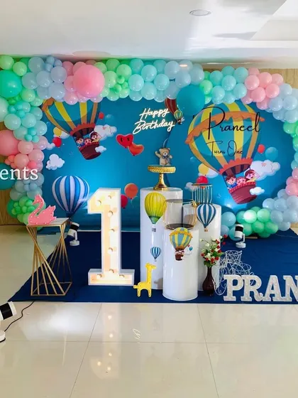 This hot air balloon theme is perfect for a 'flying high' first birthday party. The wide backdrop creates an immersive sky scene, complemented by a pastel balloon garland, marquee number, and themed props.