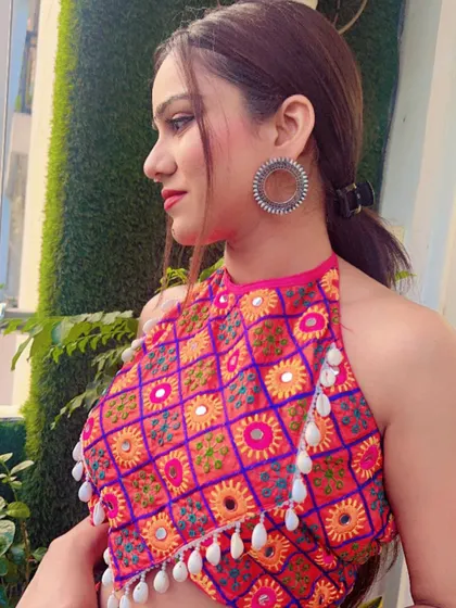 A close-up of a vibrant pink and orange halter neck blouse with mirror work and shell tassels, a modern twist on Garba wear.