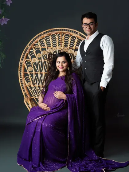 A regal portrait of the couple. The mom-to-be is seated on a peacock chair in a stunning purple saree, creating a look of pure elegance.