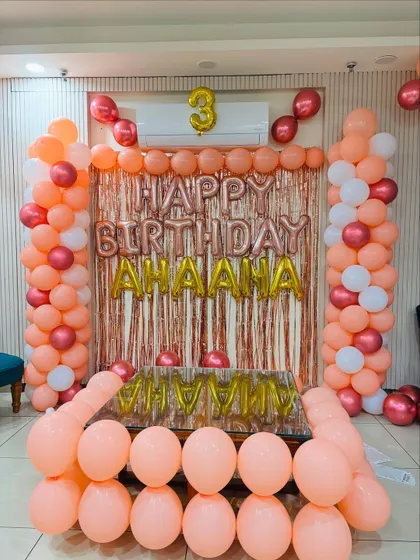 A beautiful 3rd birthday setup in peach, rose gold, and white. The decor includes balloon pillars and a tinsel backdrop with the birthday girl's name.