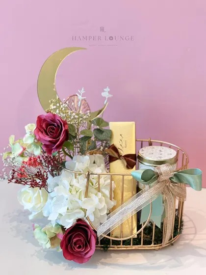 A beautiful Eid hamper arranged in a golden wire basket. It includes a scented candle, a box of chocolates, and a floral arrangement featuring red and white roses, all centered around a decorative golden moon.