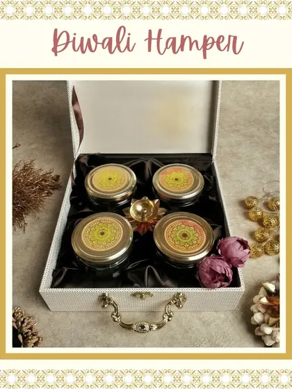 A sophisticated Diwali gift featuring a white leather-finish trunk box. Inside are four jars of dry fruits, a lotus diya, and decorative flowers, all arranged on a satin cloth.
