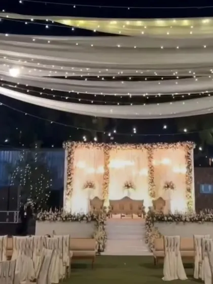 A wide view of an outdoor reception setup at night. The elegant white stage is complemented by a ceiling of fairy lights and drapes, creating a magical ambiance.