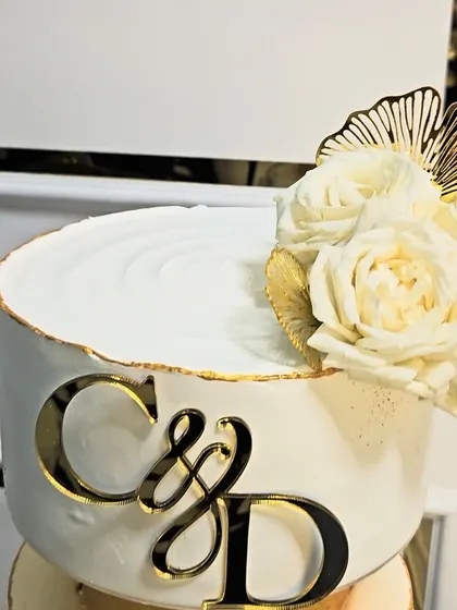 A close-up of the elegant two-tier engagement cake, showing the custom initial topper and the delicate arrangement of fresh white roses and golden fans.