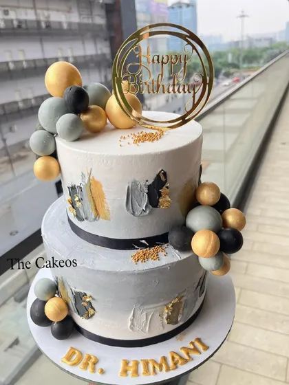 A chic and modern two-tier birthday cake for a doctor. The design features abstract brushstrokes in grey, black, and gold, with balloon-like chocolate spheres for a celebratory feel.